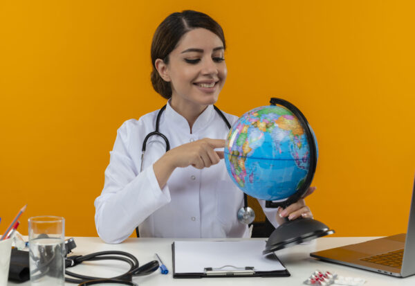 smiling young female doctor wearing medical robe and stethoscope sitting at desk with medical tools and laptop holding looking at and pointing finger to globe isolated on yellow background