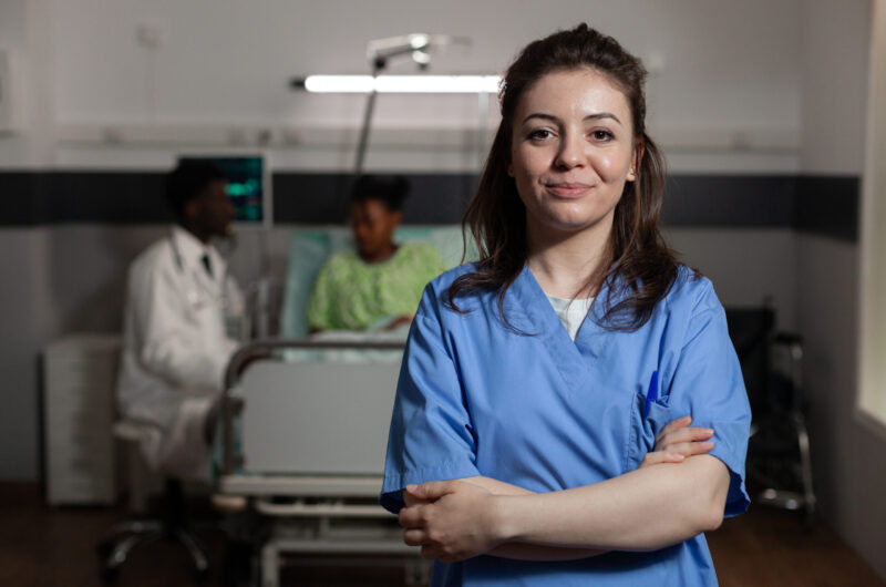 Portrait of specialist physician assistant standing in hospital ward during medical consultation. In background doctor consulting sick patient discussing healthcare treatment. Medicine services